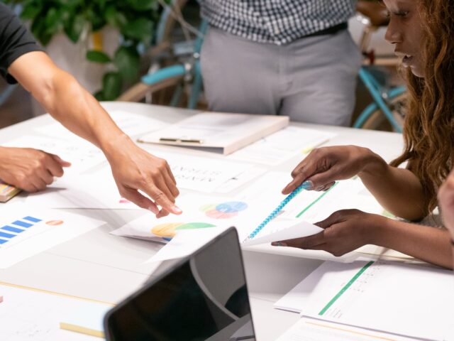 Three people with different skin tones gathered around a table with different reports printed out. They're reviewing the data and conducting survey analysis together. 